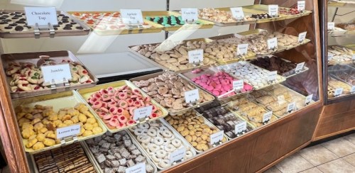 A display case filled with various colorful pastries and cookies, labeled with names, in a bakery setting.