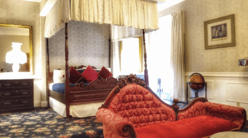 A luxurious bedroom featuring a four-poster bed, ornate red couch, and elegant decor with patterned wallpaper.