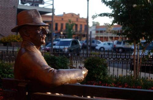 A bronze statue of a man in a hat sitting on a bench, with a street and buildings in the background.