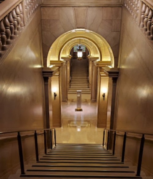 A grand staircase leads down to an elegant hallway with arched ceilings and warm lighting.