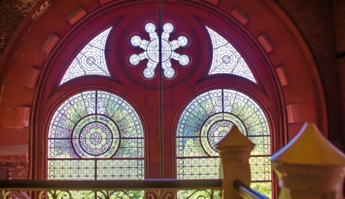Stained glass windows with intricate designs, framed by a red arch, overlooking a green landscape.