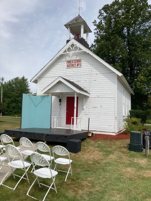 A small, white historic schoolhouse with a red door, surrounded by green grass and empty white chairs set up for an event.