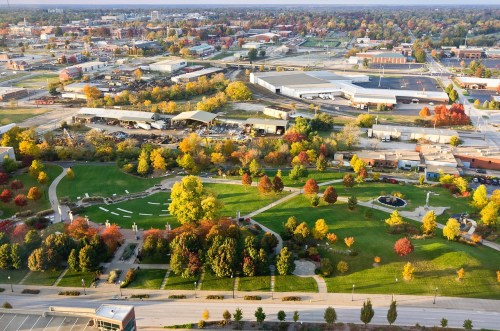 Aerial view of a colorful autumn park with trees, pathways, and nearby industrial buildings in a city landscape.