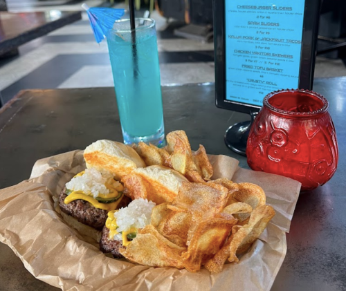 A plate with two cheeseburger sliders topped with onions and pickles, served with crispy chips and colorful drinks.