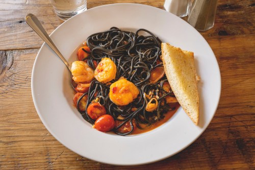 A plate of black spaghetti with shrimp, cherry tomatoes, and a slice of toasted bread on a wooden table.