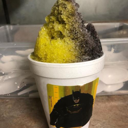 A cup of colorful shaved ice with yellow and black flavors, featuring a Batman sticker on the side.