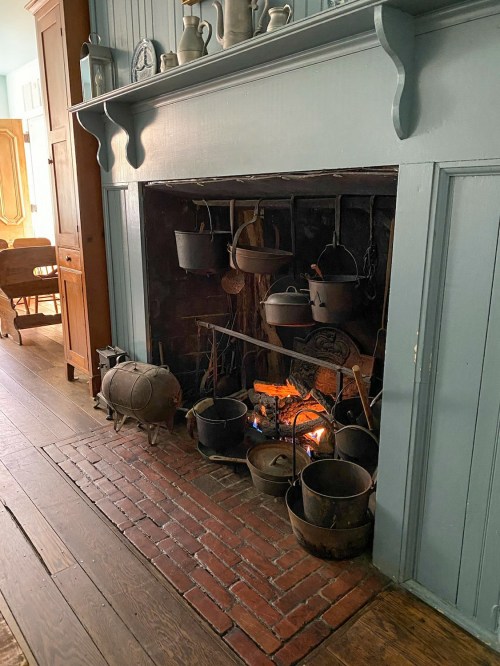 A cozy fireplace with various pots and a glowing fire, set in a rustic room with wooden floors and blue walls.