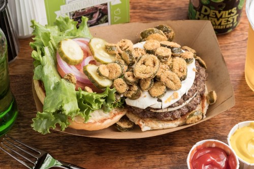 A burger with lettuce, tomato, pickles, and fried jalapeños on a wooden table, accompanied by condiments.