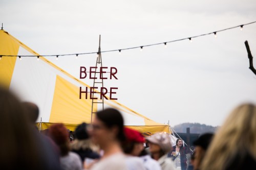 A crowd at an outdoor event with a tent displaying a sign that says "BEER HERE" and string lights overhead.
