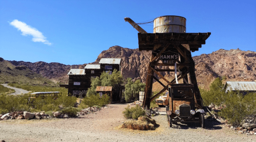 A rustic old mining town with wooden buildings, a water tower, and a vintage truck against a mountainous backdrop.