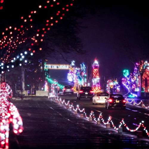 Colorful holiday lights illuminate a street at night, with cars driving past festive displays and decorations.