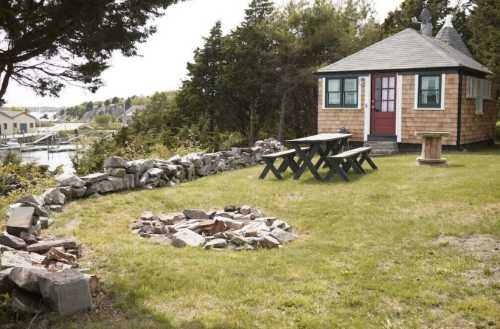 A cozy cottage with a red door, surrounded by a stone wall, picnic table, and a fire pit in a grassy area.