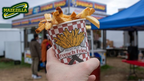 A hand holds a cup of fresh hot French fries in front of a food stall at an outdoor event.