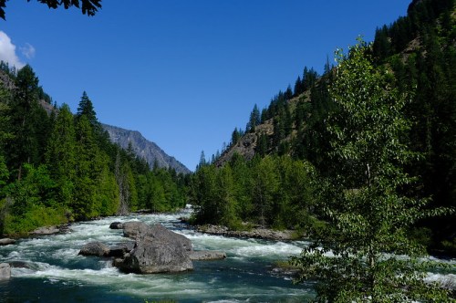 A serene river flows through a lush green landscape, surrounded by mountains and clear blue skies.