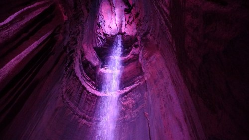 A vibrant purple-lit cave with a waterfall cascading down from the ceiling, surrounded by textured rock walls.