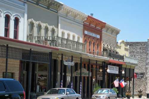 Historic buildings line a street, featuring shops and balconies, with cars parked in front under a clear blue sky.