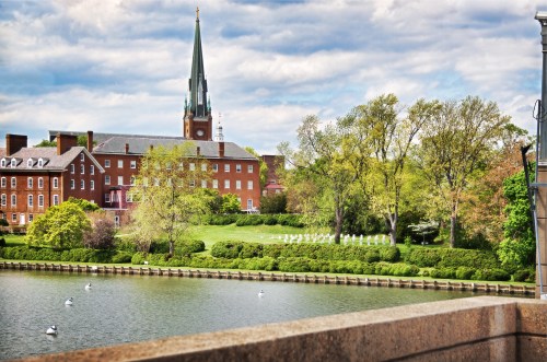 A scenic view of a lake with greenery, a brick building, and a tall spire under a cloudy sky.