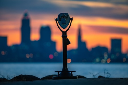 A coin-operated viewer stands in silhouette against a colorful sunset and city skyline in the background.
