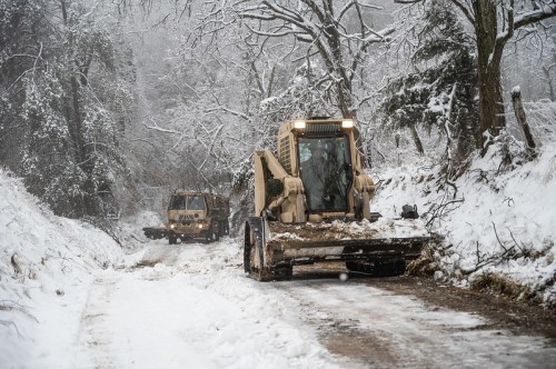 Two snow-covered construction vehicles clear a snowy road surrounded by trees in a winter landscape.