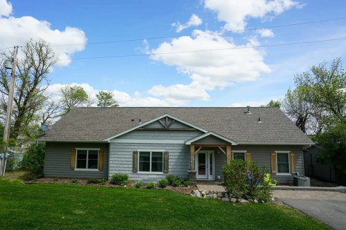 A gray single-story house with a front porch, surrounded by greenery and a clear blue sky with clouds.