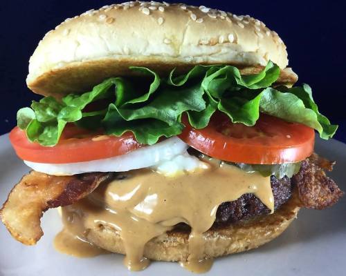 A close-up of a juicy burger with lettuce, tomato, onion, bacon, and a creamy sauce on a sesame seed bun.