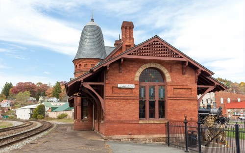 Historic red brick train station with a distinctive tower, surrounded by trees and a railway track.