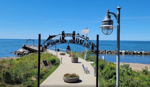 A scenic view of a waterfront path with a decorative arch, benches, and a clear blue sky over the water.