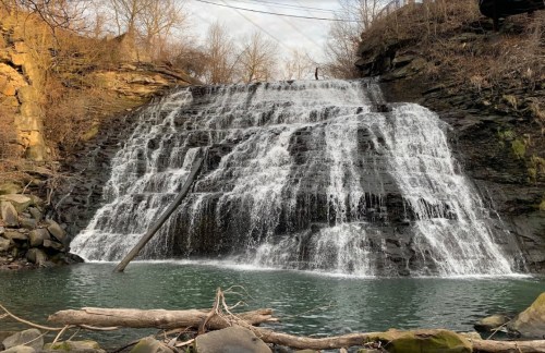 A cascading waterfall flows over rocky cliffs into a serene pool, surrounded by bare trees and natural scenery.