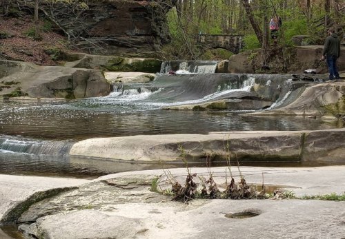 A serene waterfall cascades over smooth rocks, surrounded by trees and a calm pool of water. Two people are in the background.