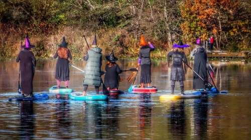 A group of people in witch costumes paddleboarding on a calm lake, surrounded by autumn foliage.