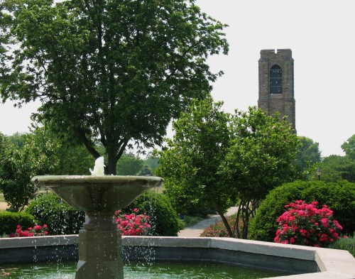 A fountain in a garden with blooming flowers, framed by trees and a distant clock tower.