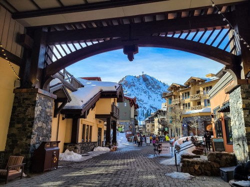 A snowy mountain view framed by a wooden archway, with shops and people in a cozy alpine village.