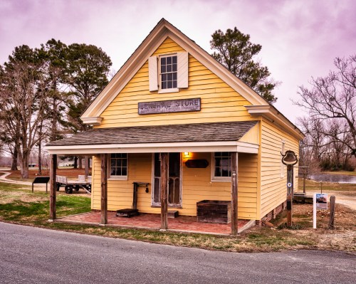A charming yellow general store with a sloped roof, surrounded by trees and a grassy area, under a purple sky.