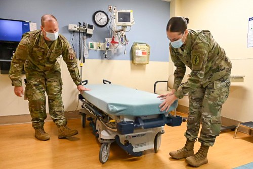 Two military personnel in camouflage uniforms adjust a hospital bed in a medical facility. Both are wearing masks.