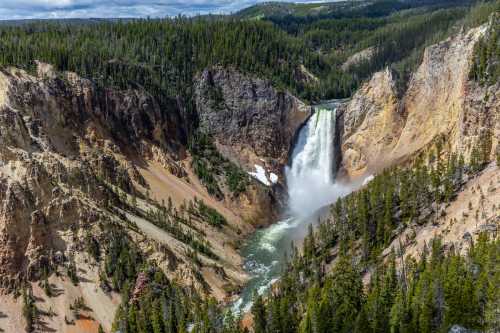 A stunning waterfall cascades down rocky cliffs, surrounded by lush green forests and mountains under a blue sky.