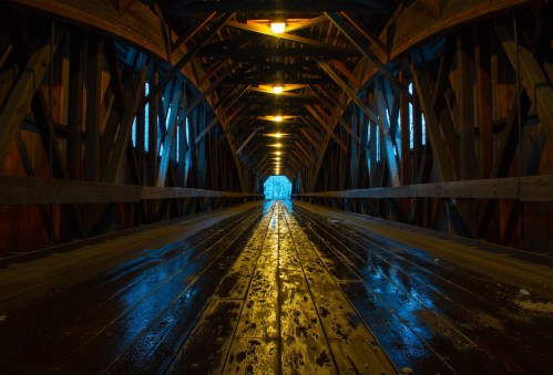 A dimly lit covered bridge interior with wooden beams and a wet floor, leading to a bright exit at the far end.