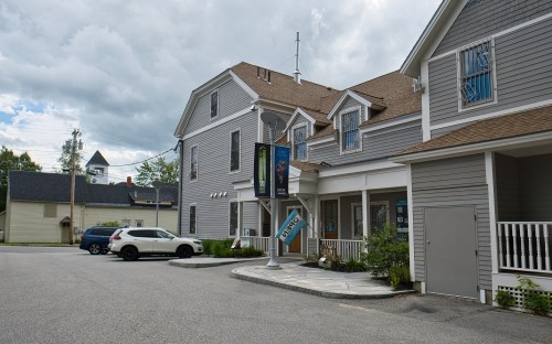 A gray building with a sign, parked cars, and cloudy skies in the background.