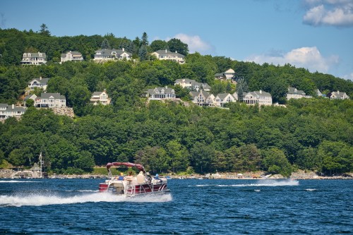 A boat speeds across a lake with lush green hills and houses in the background under a blue sky.