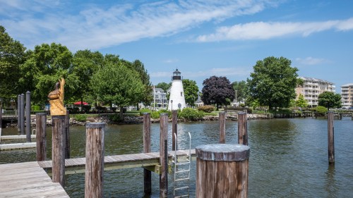 A scenic view of a lighthouse by the water, surrounded by trees and buildings, with wooden docks in the foreground.