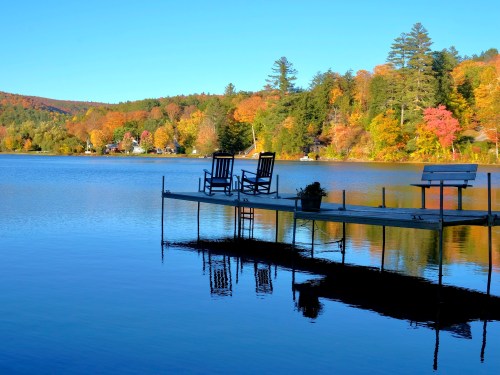 A serene lake scene with two rocking chairs on a dock, surrounded by colorful autumn foliage and clear blue skies.