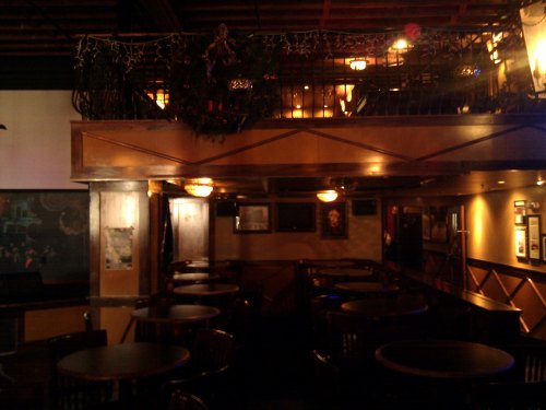 A cozy bar interior with wooden tables, dim lighting, and a balcony adorned with decorations.