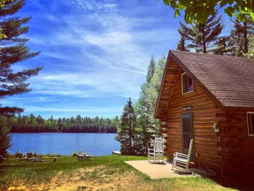 A cozy log cabin by a serene lake, surrounded by lush trees and a clear blue sky. Rocking chairs on the porch invite relaxation.