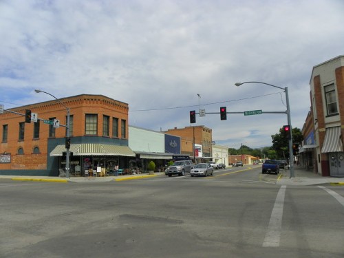 A street view of a small town intersection with brick buildings, traffic lights, and parked cars under a cloudy sky.