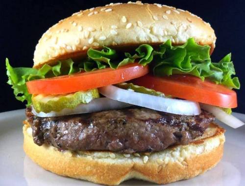 A juicy hamburger with lettuce, tomato, pickles, and onion, served in a sesame seed bun on a white plate.