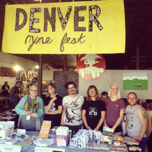 A group of six people stands behind a table at the Denver Zine Fest, with a large banner overhead.