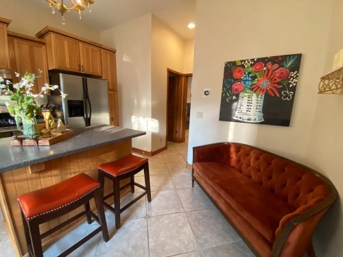 A cozy kitchen with wooden cabinets, a gray countertop, two bar stools, and a vibrant floral painting on the wall.