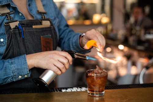 A bartender uses a torch to flame an orange peel over a cocktail glass filled with ice.