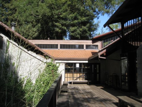A wooden deck leads to a modern building surrounded by trees and greenery under a clear blue sky.