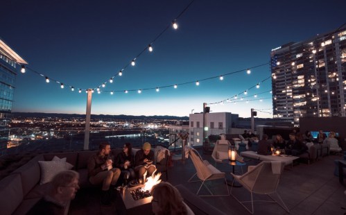 A rooftop lounge at dusk, with string lights, a fire pit, and people enjoying the view of the city skyline.
