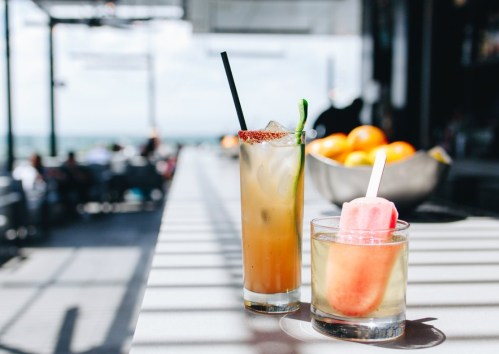Two refreshing drinks on a table: a cocktail with a straw and a popsicle in a glass, with a bright outdoor setting.